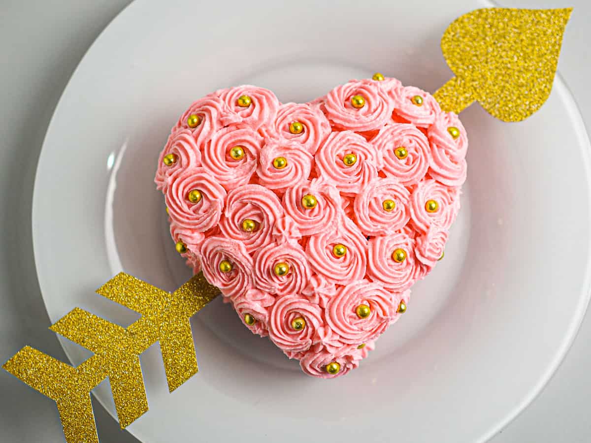 A pink buttercream rossette frosted heart shaped cake with a gold paper arrow through it.