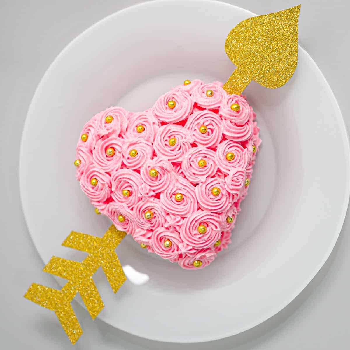 A pink buttercream rossette frosted heart shaped cake with a gold paper arrow through it.