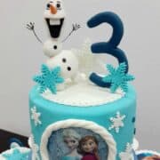3D Olaf cake topper on a light blue round cake decorated with Frozen theme.
