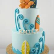 A two tier beach themed cake in blue, orange and yellow, with orange gum paste hibiscus and fondant surfboards.
