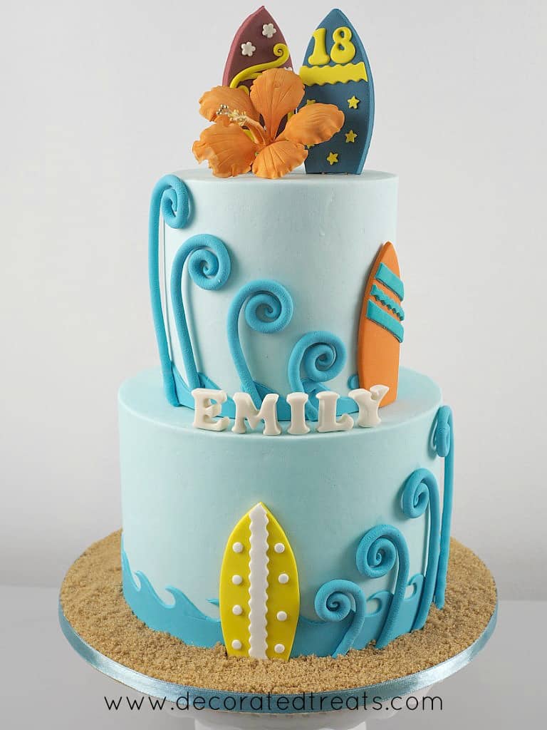 A two tier beach themed cake in blue, orange and yellow, with orange gum paste hibiscus and fondant surfboards