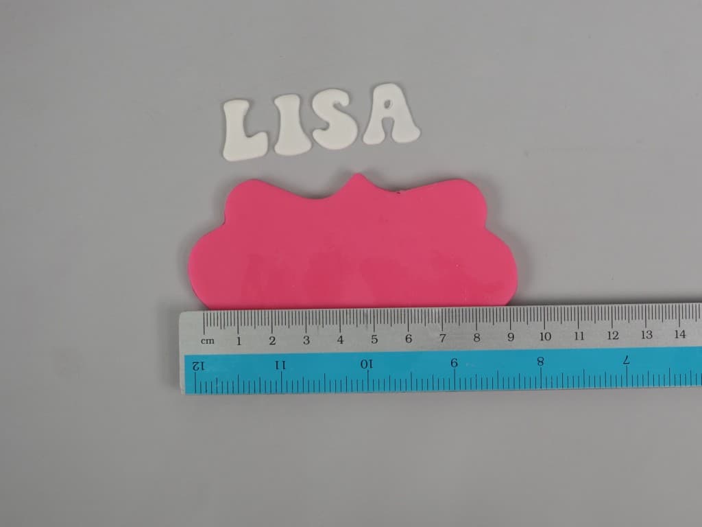 A pink fondant plaque with a ruler on it. On the side are cake lettering L, I, S and A.
