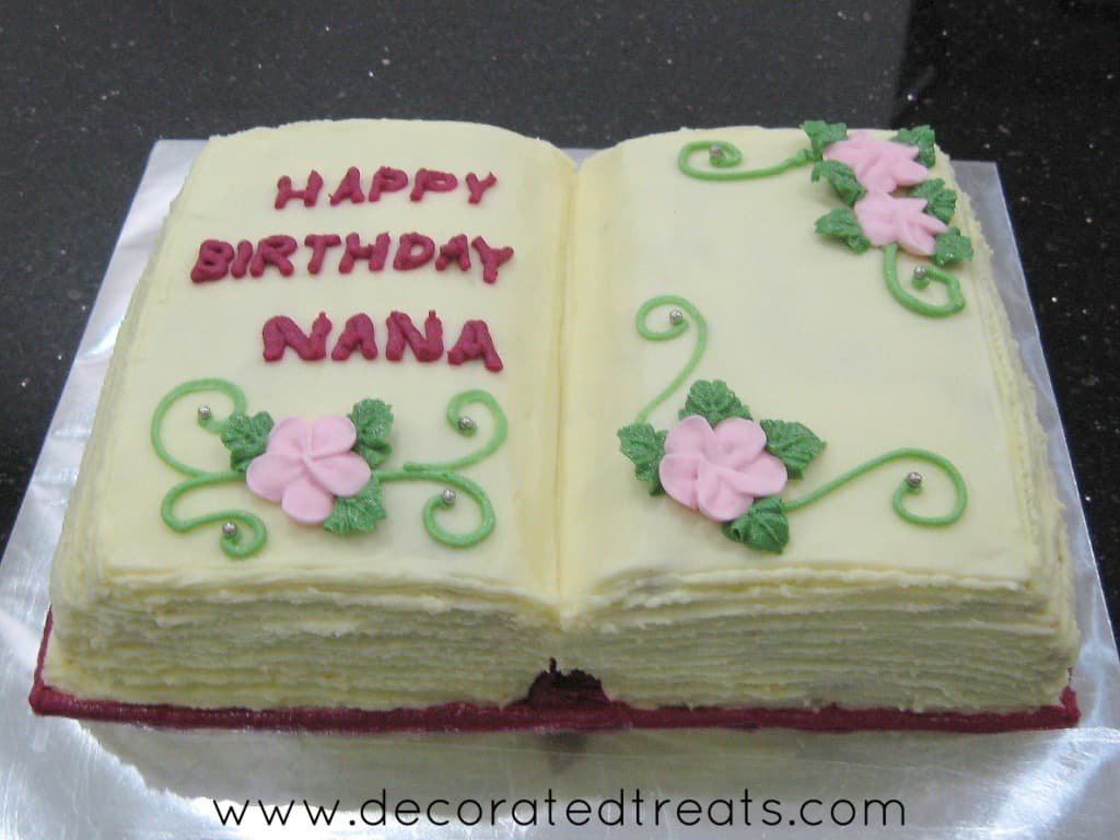A cake in the shape of an open book with pink flowers and green leaves and scrolls