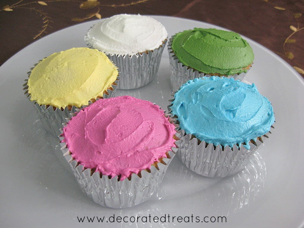 5 cupcakes on a plate. Each covered in blue, yellow, white, green and pink icing