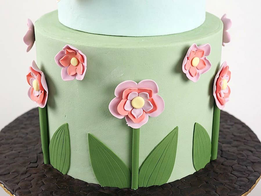 The bottom tier of a cake decorated with pink and purple flowers, leaves and stems.