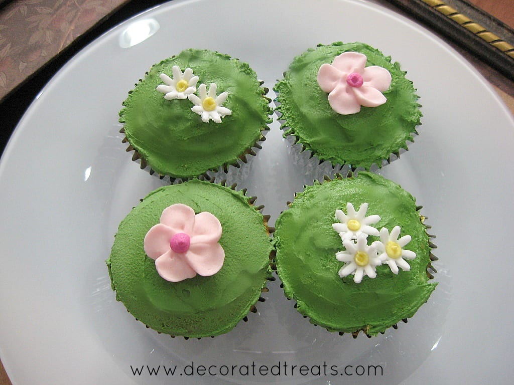 4 cupcakes on a white plate, 2 covered in white icing and pink flowers while the other 2 covered in green icing and decorated with fondant daisies.