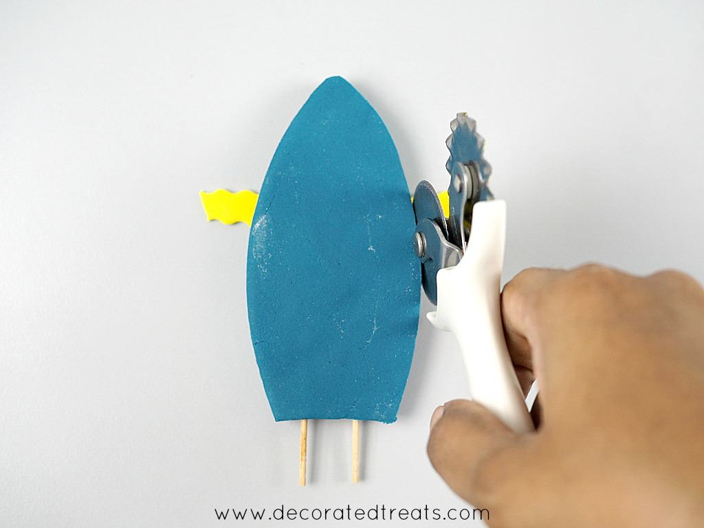 Cutting the excess of a yellow fondant strip on a blue fondant piece with a pizza cutter