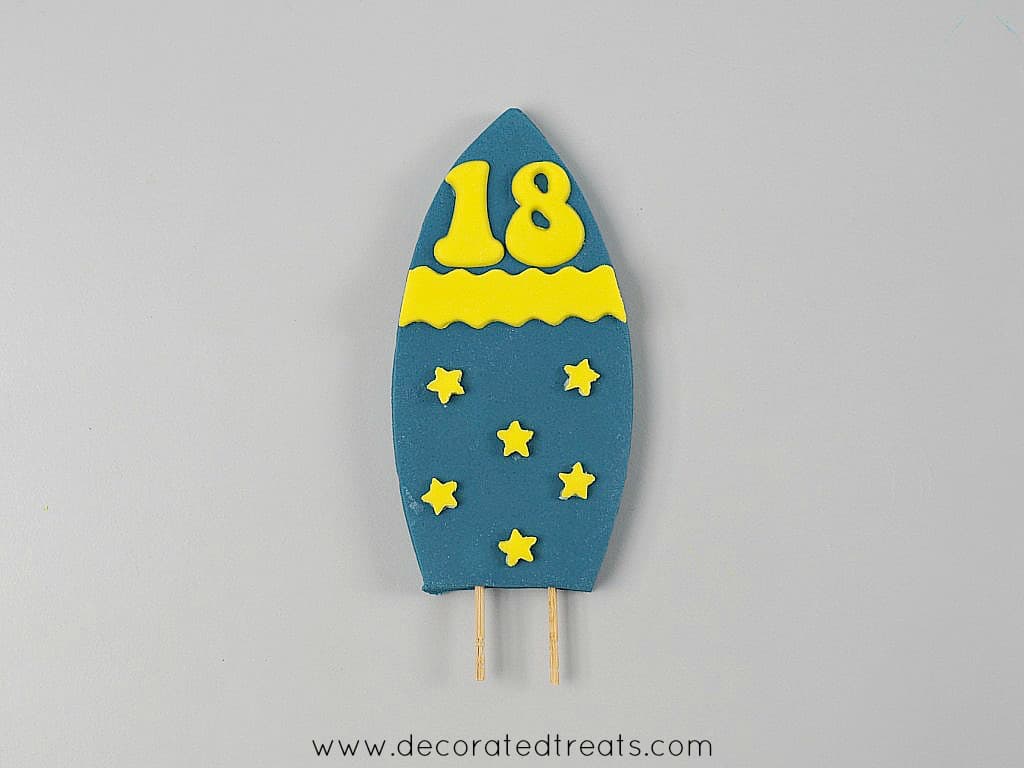Dark blue fondant surf board with toothpicks attached, decorated with yellow stars, a horizontal yellow strip and number 18