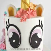 A two tier white unicorn cake with 2 large fondant eyes on the top tier.