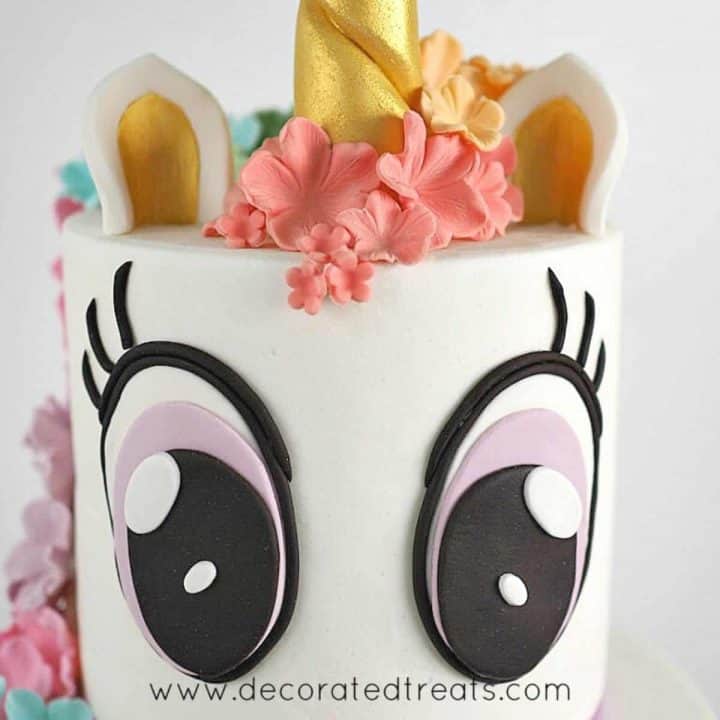 A two tier white unicorn cake with 2 large fondant eyes on the top tier