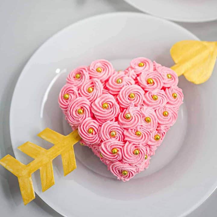 A heart shaped cake decorated with pink buttercream rossettes and a gum paste arrow going through it