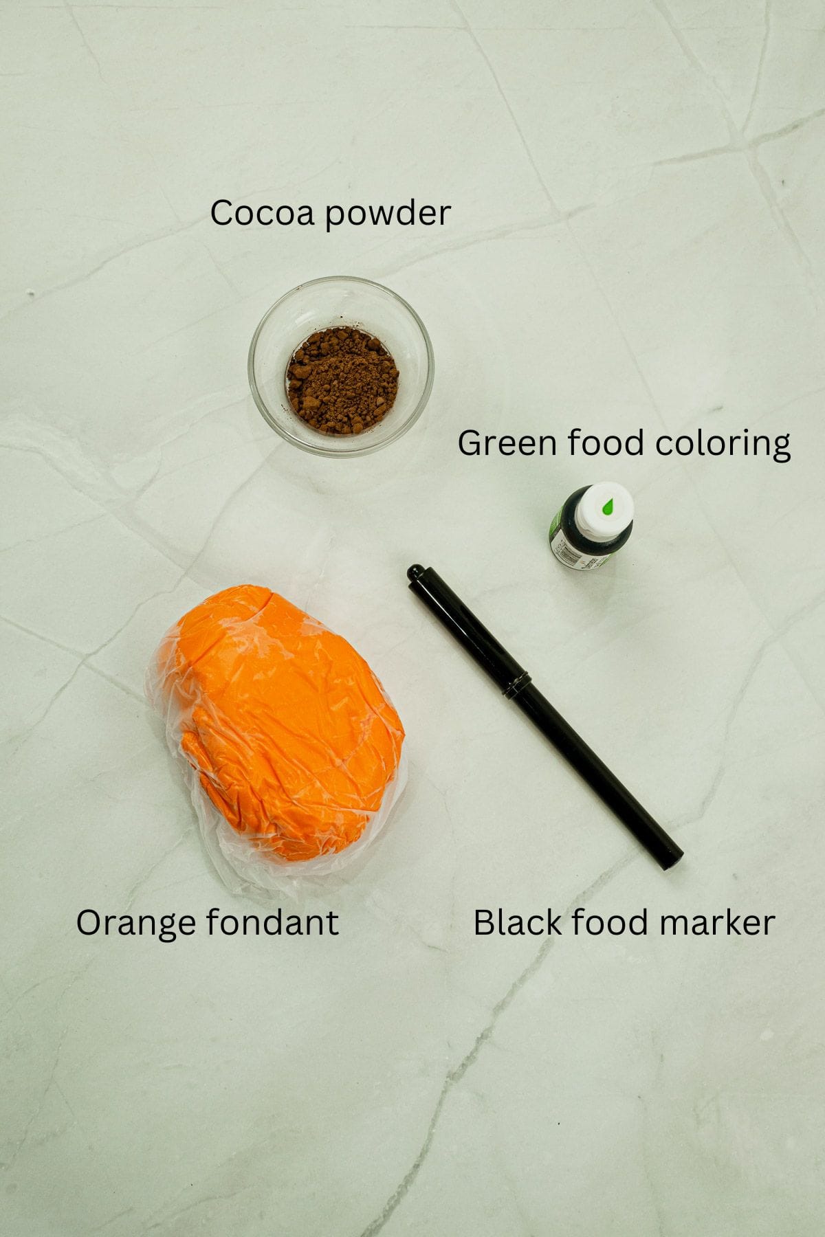 Cocoa powder, black food marker, green food coloring and orange fondant against a marble background.