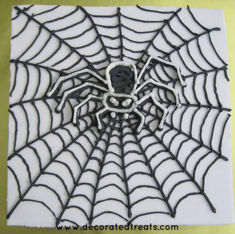 A white square cake with a royal icing spider web and a black piped spider