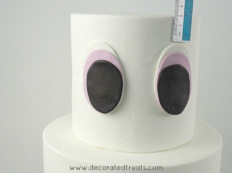 Using a ruler to measure the spacing from the top of the cake to the fondant eyes on the top tier cake.