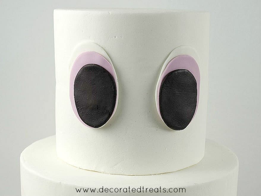 A two tier white cake with 2 large fondant eyes on the top tier.