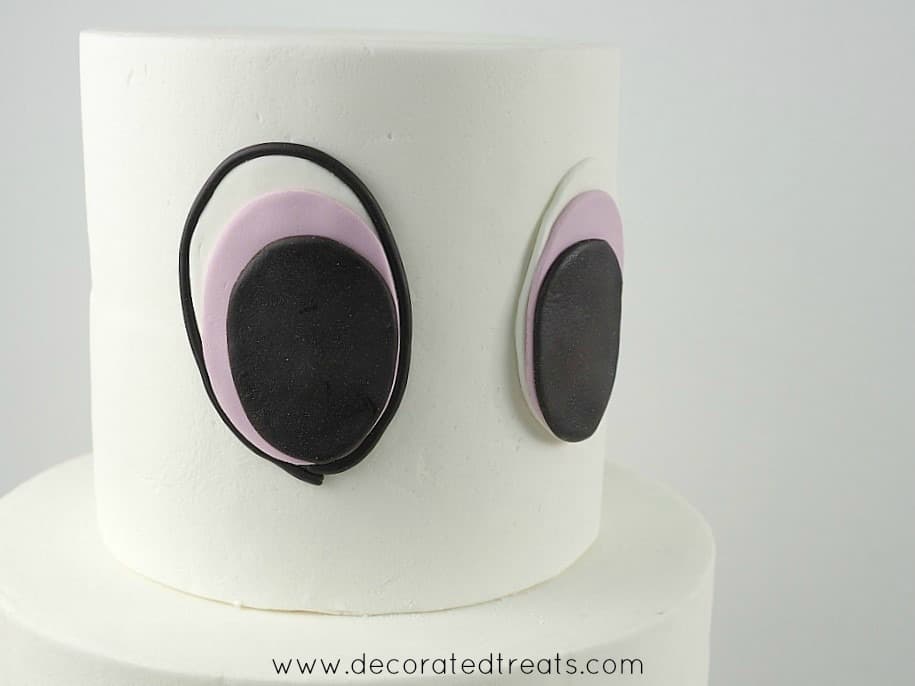 A two tier white cake with 2 large fondant eyes on the top tier