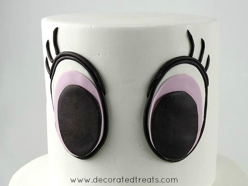 A two tier white cake with 2 large fondant eyes on the top tier.