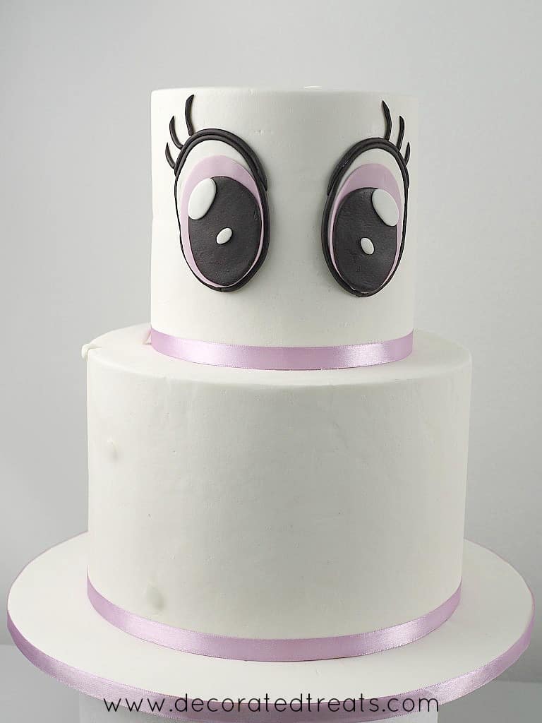 A two tier white cake with 2 large fondant eyes on the top tier