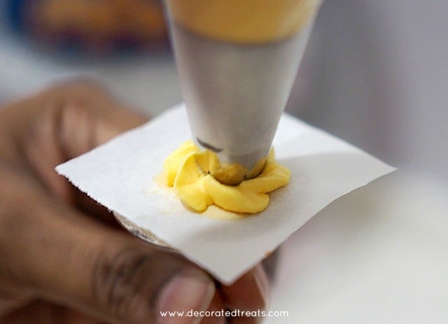 Piping an yellow buttercream flower onto a parchment square on a flower nail