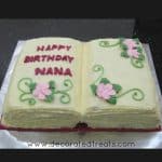 A cake in the shape of an open book with pink flowers and green leaves and scrolls.