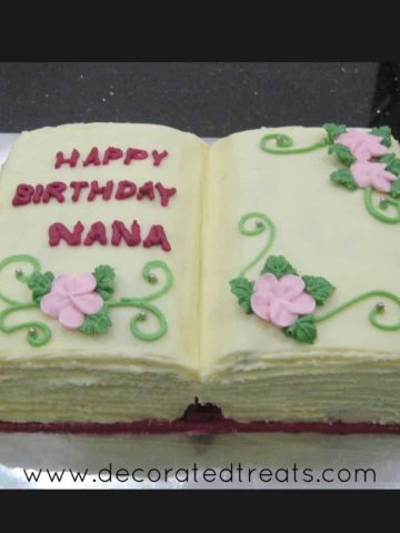 A cake in the shape of an open book with pink flowers and green leaves and scrolls.