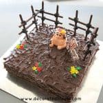 A square cake covered in ganache with a fondant pig topper and cookie sticks fencing.