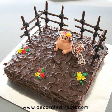 A square cake covered in ganache with a fondant pig topper and cookie sticks fencing