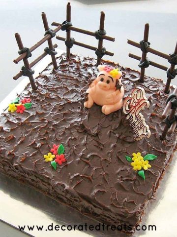 A square cake covered in ganache with a fondant pig topper and cookie sticks fencing