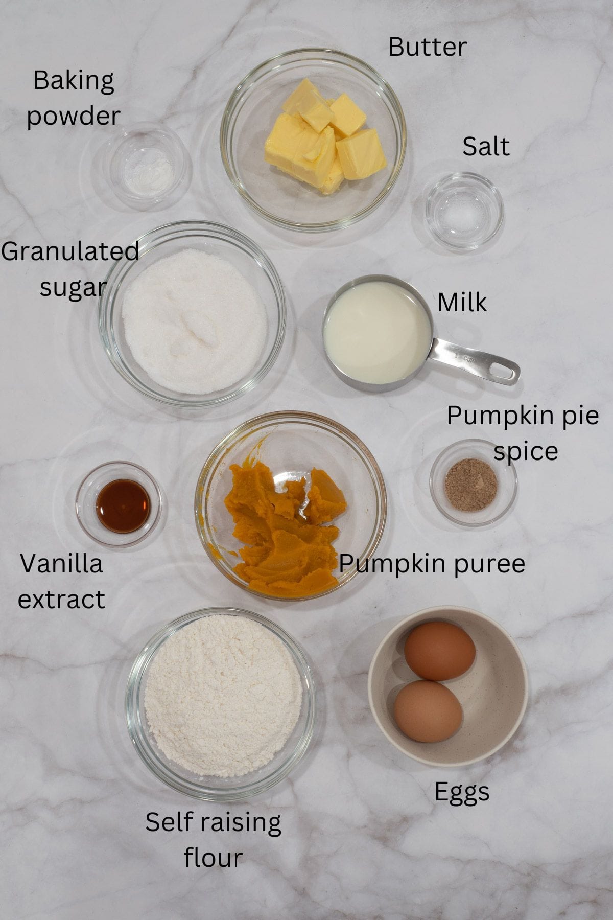 Self raising flour, granulated sugar, butter, eggs, pumpkin puree, pumpkin pie spice, vanilla extract, baking powder, salt and milk against a marble background.