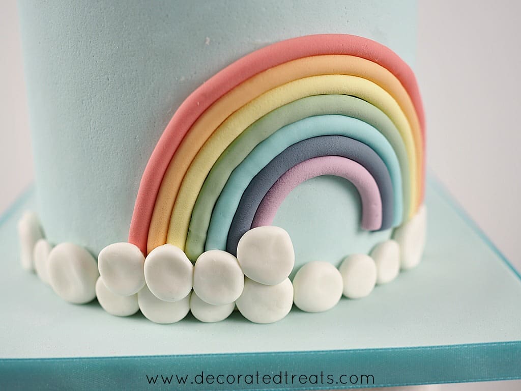 A cake decorated with fondant rainbow and a border of white fondant patches as the clouds