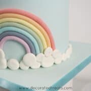 A cake decorated with fondant rainbow and a border of white fondant patches as the clouds.