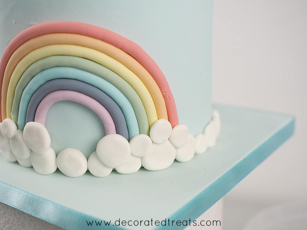 A cake decorated with fondant rainbow and a border of white fondant patches as the clouds