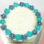 Turquoise and blue buttercream flowers arranged in a circular pattern around the top edges of a round cake covered in buttercream.