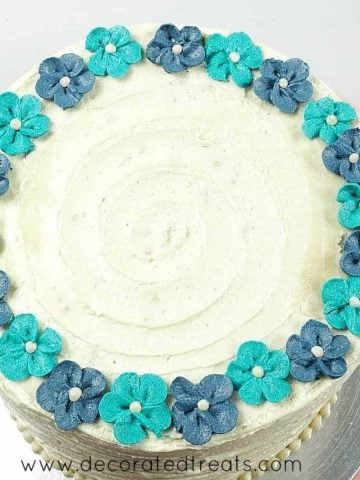 Turquoise and blue buttercream flowers arranged in a circular pattern around the top edges of a round cake covered in buttercream