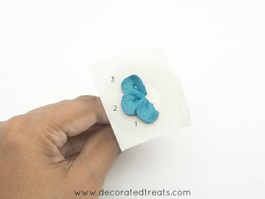 3 petals of a buttercream flower in blue piped on a parchment square held on a flower nail