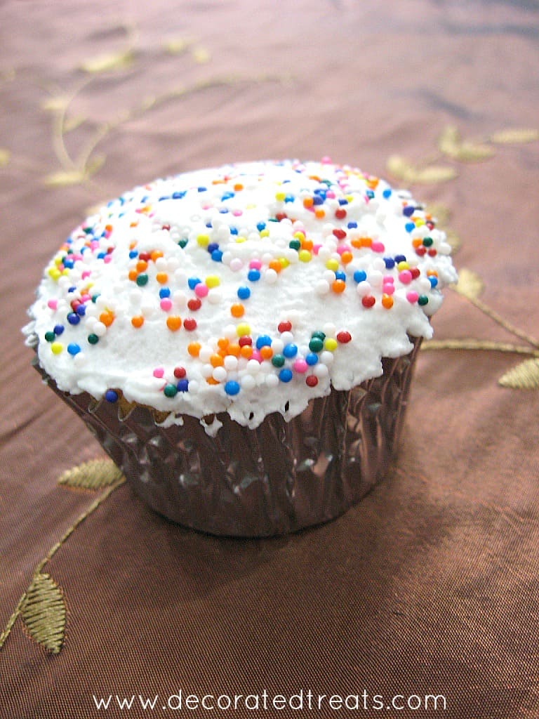 A cupcake in a silver casing, with white frosting and colorful sprinkles