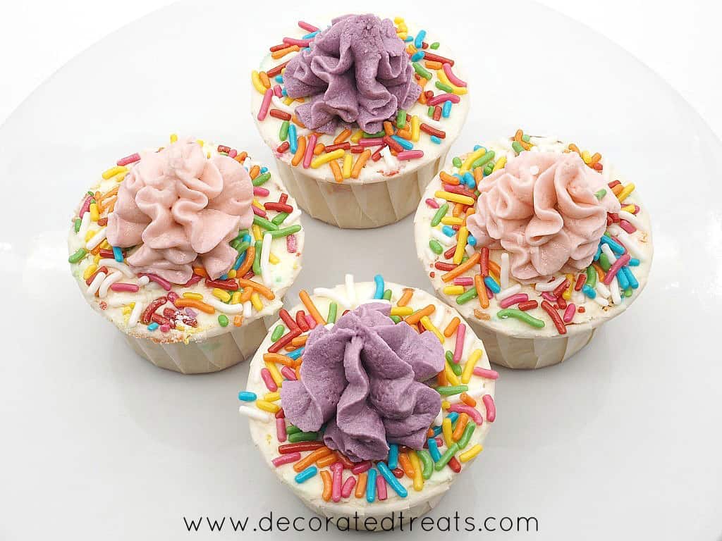 4 cupcakes on a plate, decorated with pink and purple swirl icing and colorful spinkles