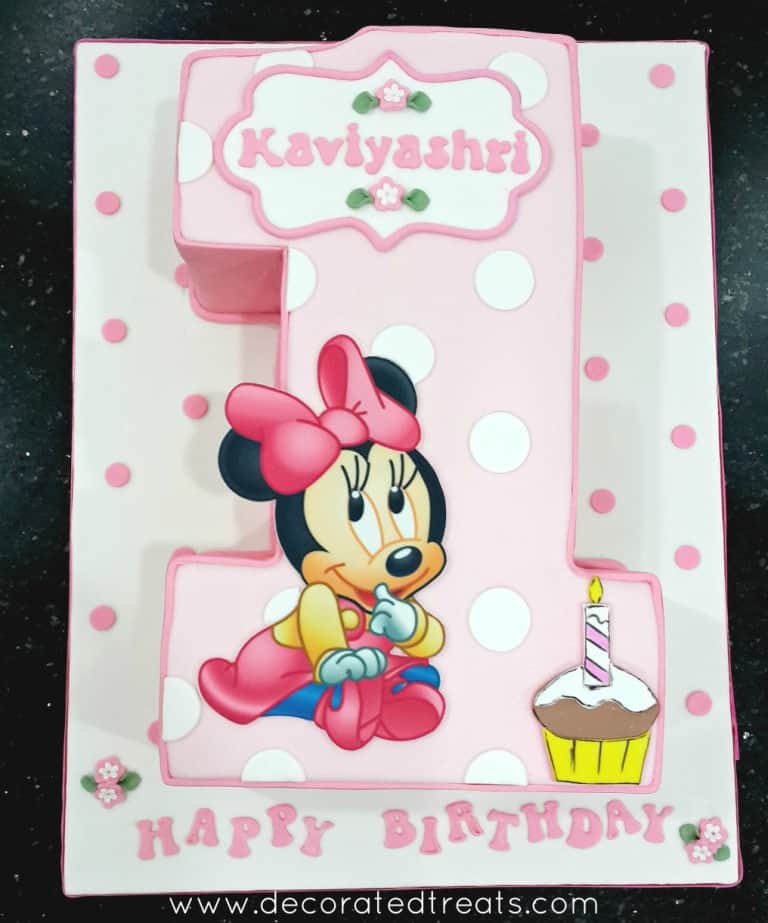 A number one shaped cake decorated with a Minnie Mouse baby image and a cup image. The cake is in pink with white polka dots while the cake board is white in pink polka dots.