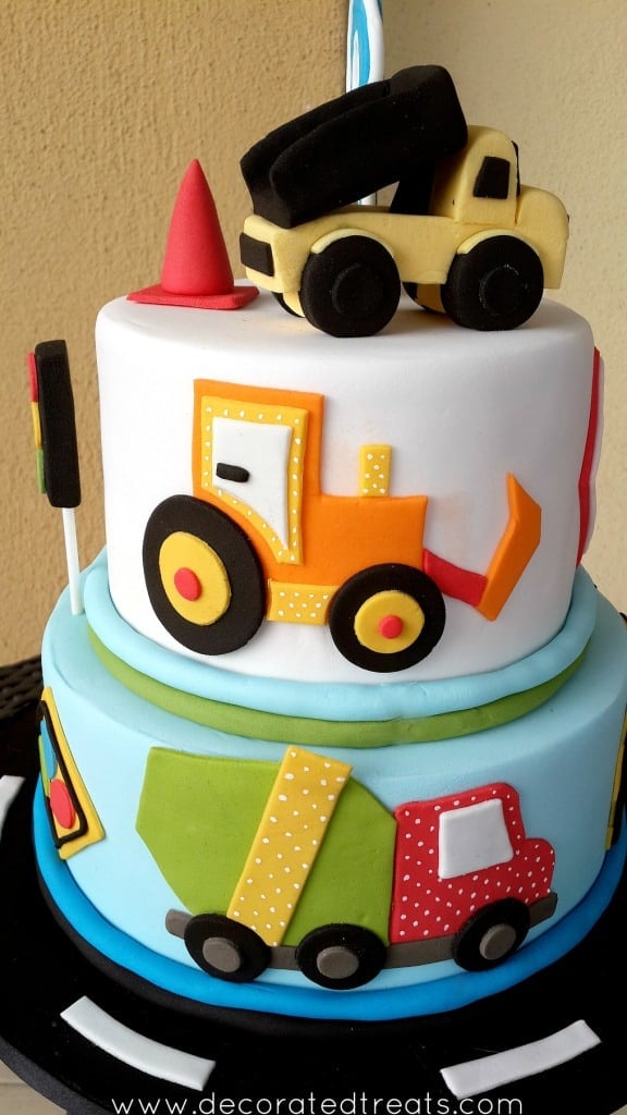 A 2 tier cake with truck topper, and the sides decorated with an orange and red and green trucks.