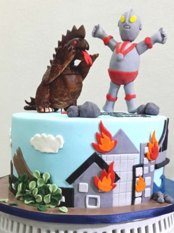 A round cake with Ultraman and monster toppers. Side of the cake is decorated with buildings on fire.
