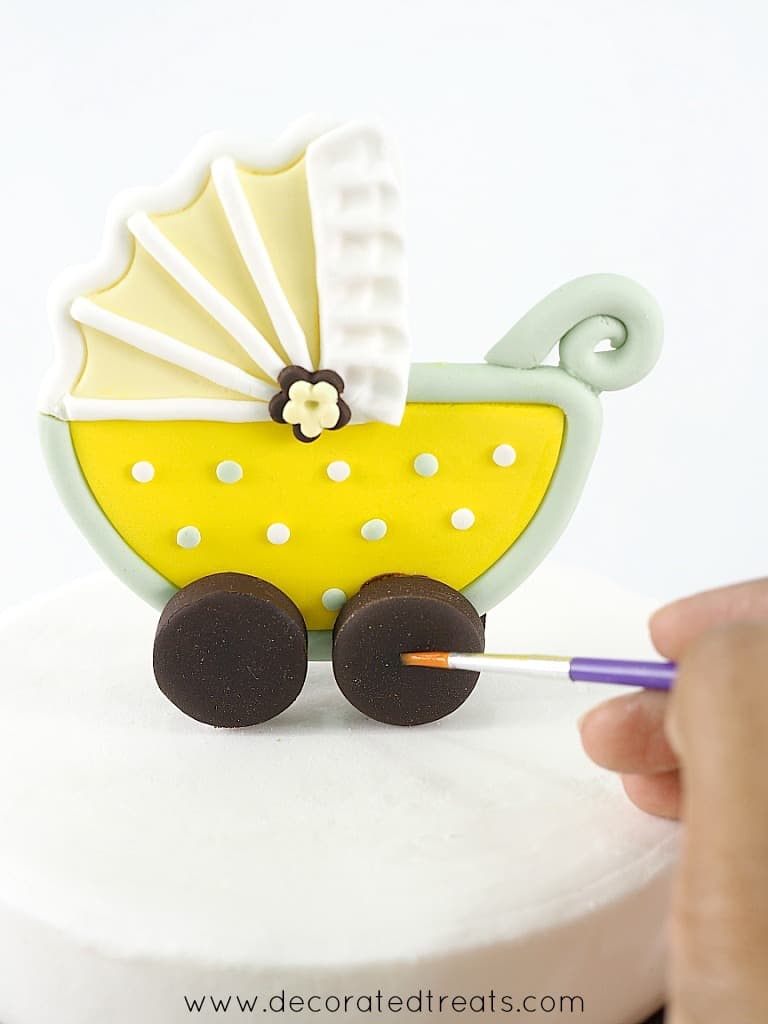 Using a brush to apply fondant glue onto the fondant stroller topper wheel for a baby shower cake.