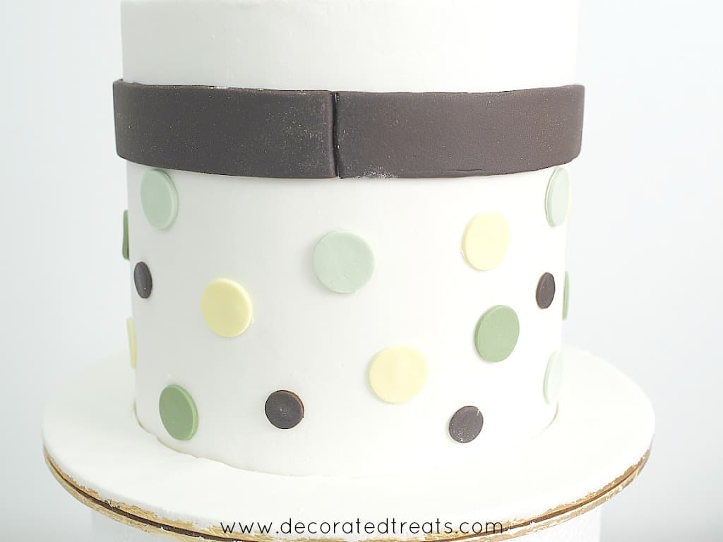A brown fondant strip around a white round baby shower cake. Cake is decorated with green, yellow and brown dots