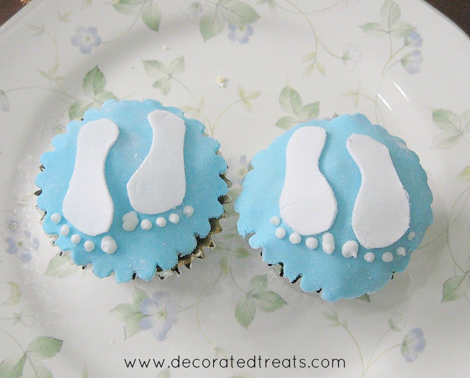2 cupcakes on a white plate, covered in blue fondant and decorated with white baby footprints