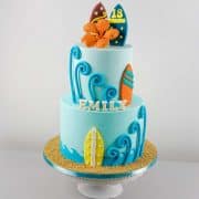 A 2 tier blue beach themed birthday cake with colorful fondant surfboards, and an orange hibiscus topper.
