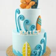 A 2 tier blue beach themed birthday cake with colorful fondant surfboards, and an orange hibiscus topper.