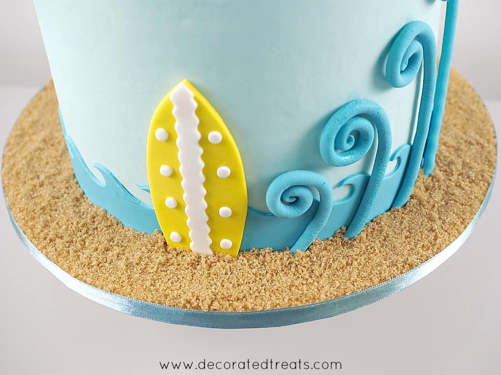 Cake board covered in brown sugar. Cake is covered in blue fondant and there is a yellow fondant surfboard deco on the side.