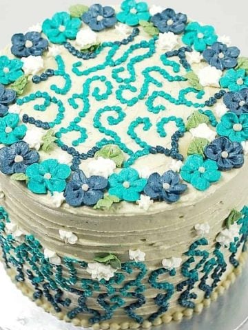 A round beige cake decorated with blue and turquoise buttercream piping and white flowers.