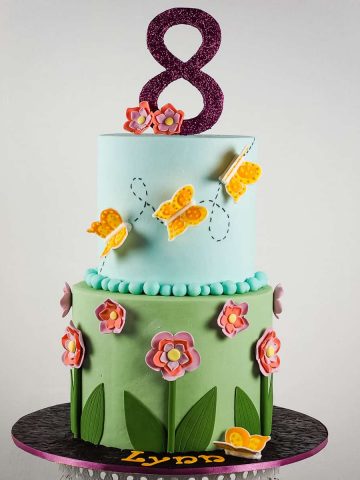 A 2 tier round cake in green and blue fondant. Bottom tier is decorated with fondant flowers and the top tier with royal icing butterflies in orange. Cake topper is a large glittery number 8.