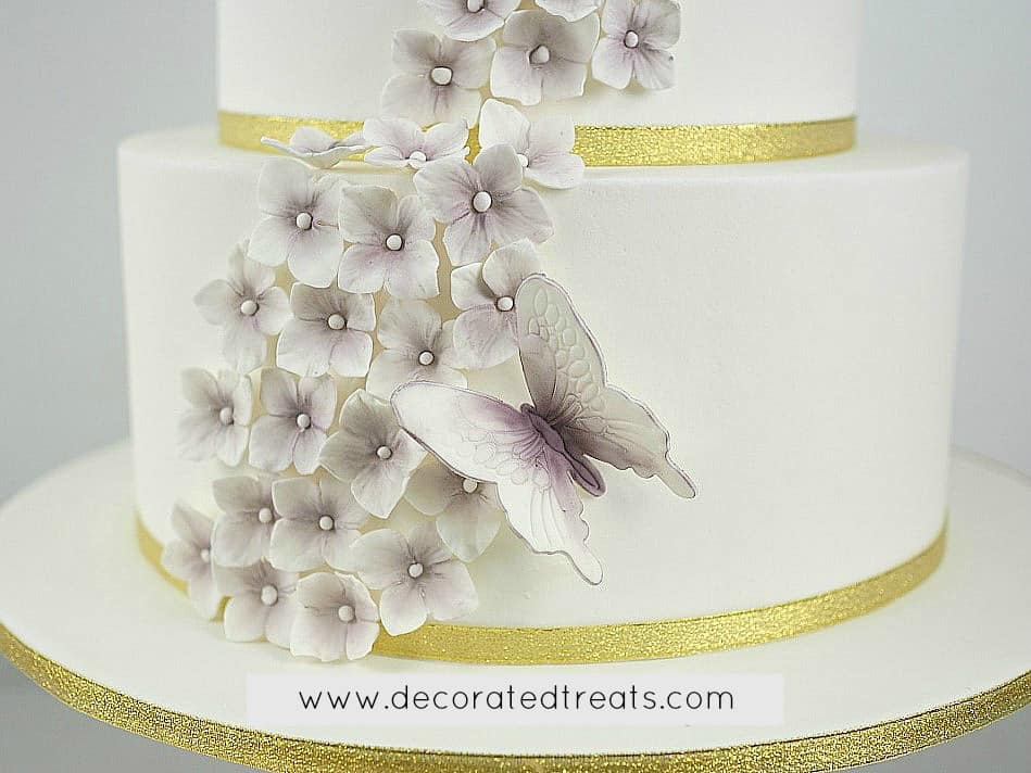 Gum paste butterflies and hydrandea in purple on a white cake with gold ribbon border