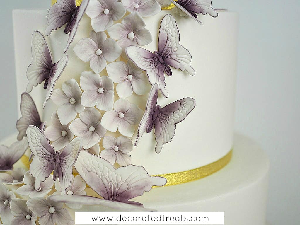 Gum paste butterflies and hydrandea in purple on a white cake with gold ribbon border